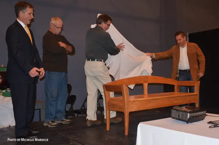 Men unveiling wooden bench on stage.
